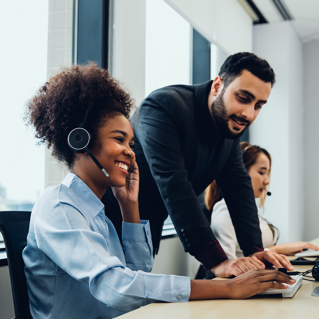 Office setting with a person wearing a headset working on a computer while another person stands nearby providing assistance.