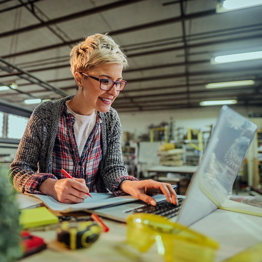 Person with short blond hair and glasses smiling while working on a laptop and sketching at a workbench in a workshop setting