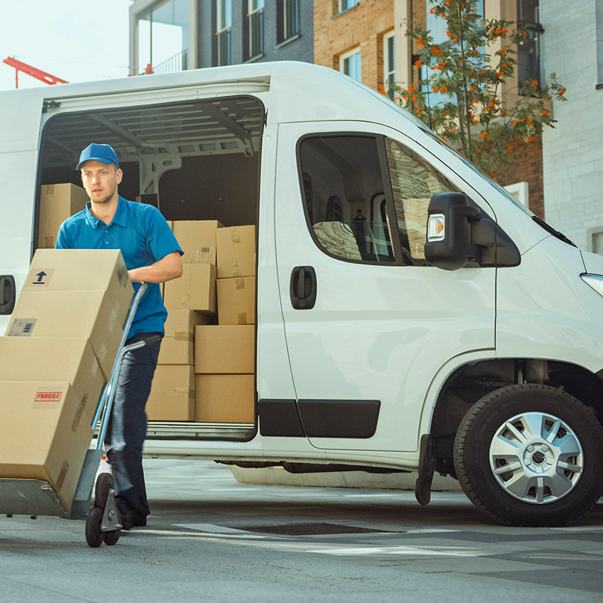 Delivery worker unloading cardboard boxes from a white cargo van using a hand truck in front of a modern building