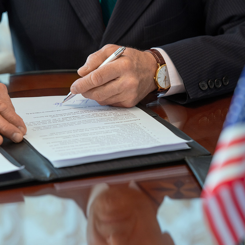 Close-up of a person in a business suit signing official documents on a wooden desk with an American flag in view.