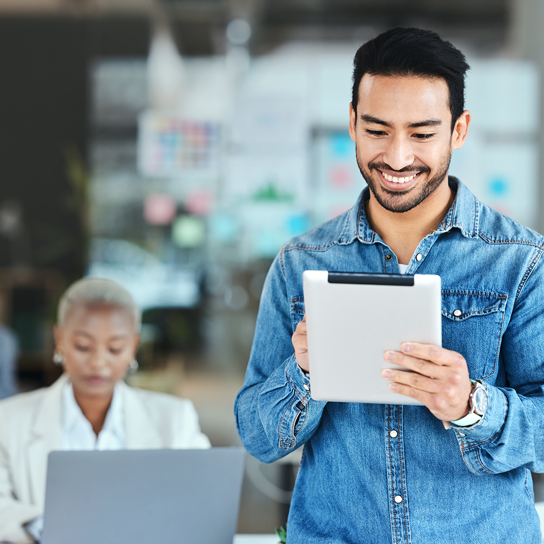 Person in a denim shirt holding a tablet while standing in a modern office, with another person working on a laptop in the background.