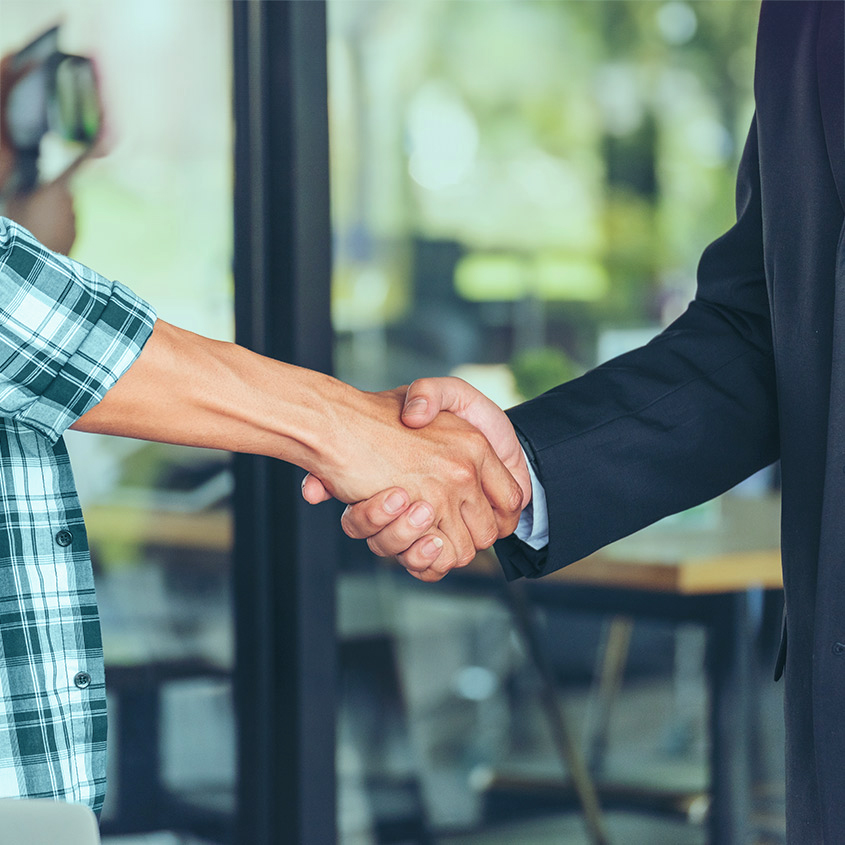 Close-up of two people shaking hands indoors, one wearing a plaid shirt and the other in a business suit.