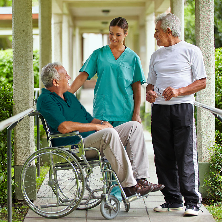 Healthcare professional standing outdoors with two older adults, one seated in a wheelchair and the other standing nearby.