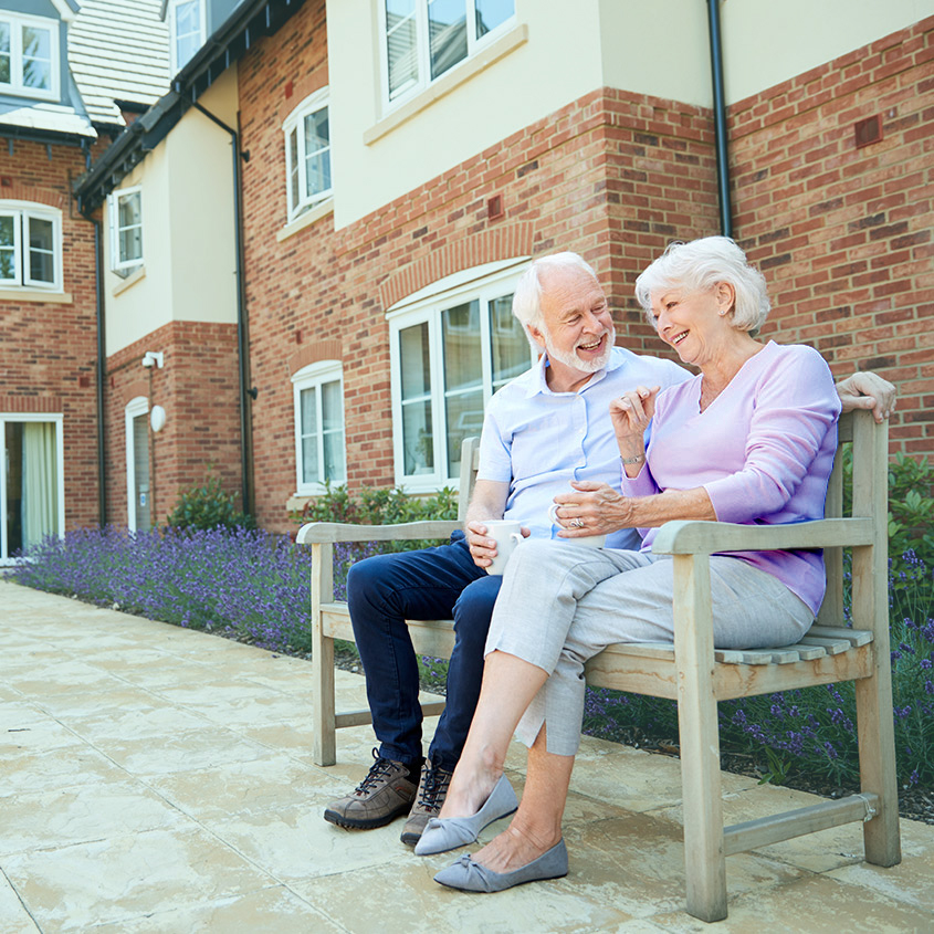 Two older adults sitting on a wooden bench outside a brick senior living facility, enjoying drinks in a landscaped courtyard.